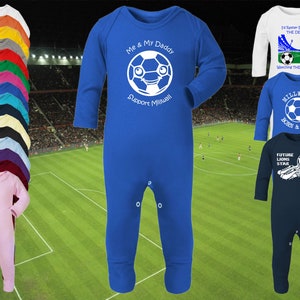 Può includere: Un body blu per neonato con testo bianco che recita "Io e il mio papà sosteniamo il Millwall" e un pallone da calcio bianco disegnato con un viso. Sullo sfondo ci sono altri tre body per neonato, uno bianco con testo blu, uno blu con testo bianco e uno blu scuro con testo bianco.
