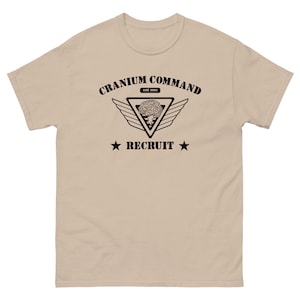 以下が含まれることがあります： ベージュ色のTシャツ。黒いグラフィックデザインが特徴です。デザインには、「Cranium Command」の文字が、脳のイメージが入った三角形の上に配置され、翼が両側に配置されています。「Recruit」の文字が下にあり、星が両側にあります。Tシャツはクルーネックです。