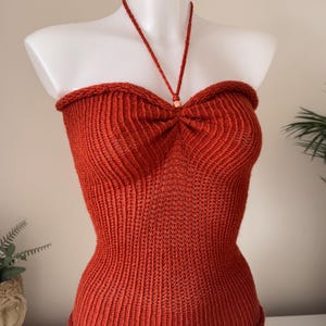 May include: A rust-coloured, ribbed knit halter top featuring a sweetheart neckline with a central gathered detail and a thin neck strap secured by a small, round, light-coloured bead. The garment has a fitted silhouette with a rolled hem at the top.