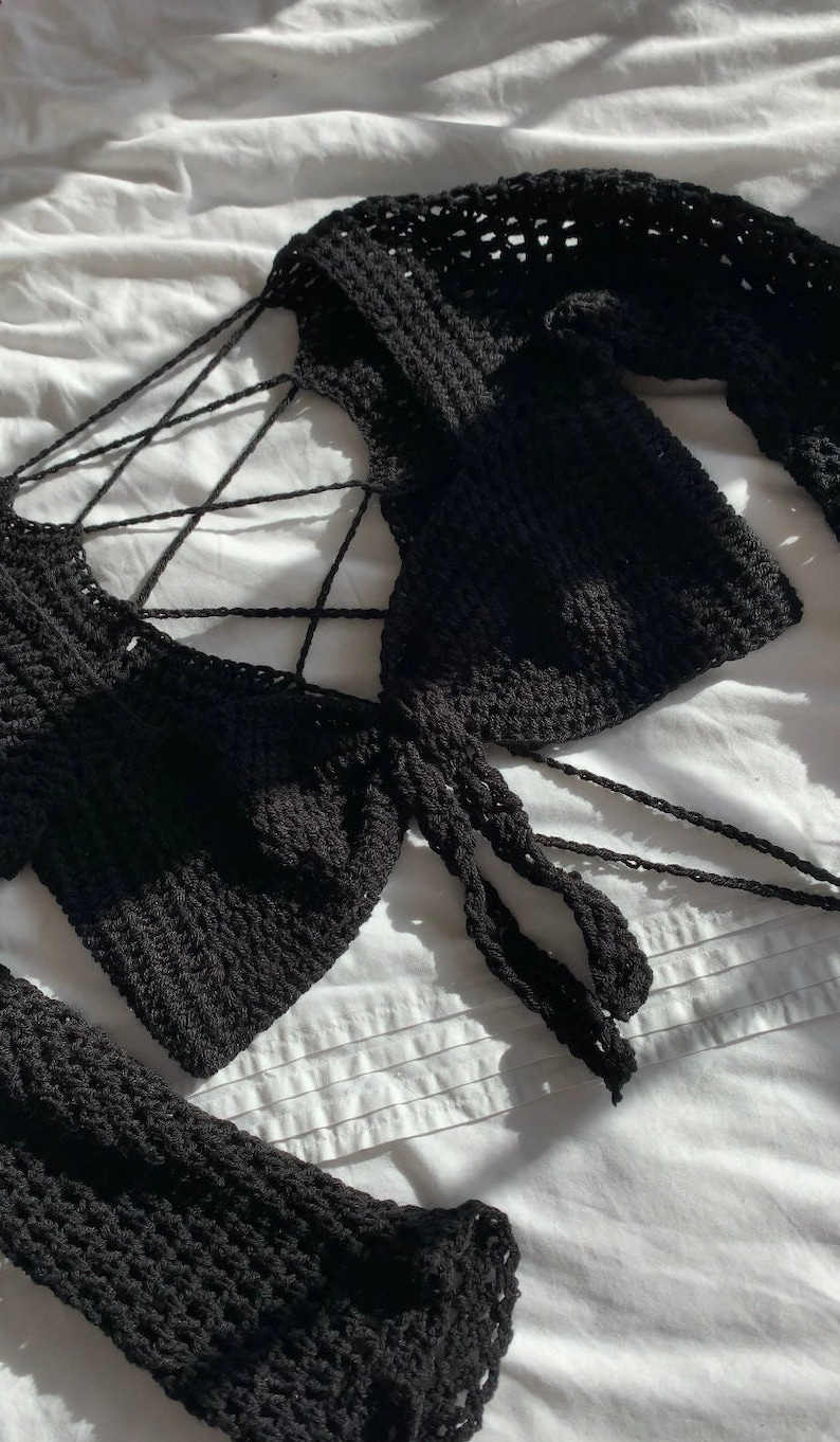 K&ouml;nnte beinhalten: Ein schwarzes H&auml;keltop mit langen &Auml;rmeln und einem Kreuz-Knoten-Detail an der Vorderseite.