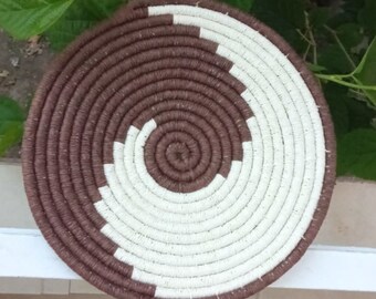 May include: A round woven basket with a brown and white spiral pattern. The basket is made of natural materials and has a rustic look.