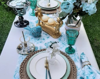May include: A table setting with a white tablecloth featuring a blue floral pattern. The centrepiece is a vase filled with blue and white flowers. Two place settings are visible, each with a white plate, green charger, and gold cutlery. The table is adorned with a rope placemat, a bird figurine, and a candle holder with two white candles.
