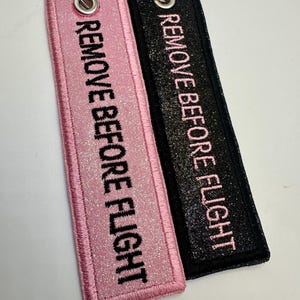 May include: Two rectangular keychains with the text "REMOVE BEFORE FLIGHT". One is pink with silver glitter, the other is black with silver glitter. Both have a silver metal ring.