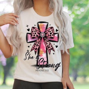 May include: White t-shirt with a pink cross design, a leopard print bow, and the text "She is Strong" and "Proverbs 31:25". The design includes black accents and small heart and cross details.