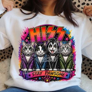 Pode incluir: Camisola branca com um gráfico colorido de quatro gatos ao estilo da banda KISS. Os gatos usam jaquetas com tachas e maquiagem, com as palavras "KISS" e "STAY PAWSOME".