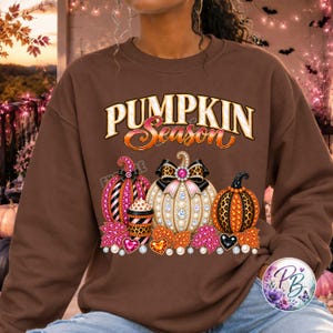 May include: A brown crewneck sweatshirt featuring the text PUMPKIN SEASON in white and orange. The graphic includes three pumpkins decorated with leopard print, pink glitter, and faux pearls, alongside a coffee cup and colorful autumn leaves.