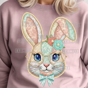 May include: A pink sweatshirt featuring a colorful patchwork bunny face with heart-shaped glasses, a bow tie, and floral accents. The bunny has large blue eyes and long ears. The design is detailed and whimsical.