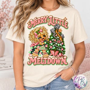 May include: A cream-colored t-shirt featuring a graphic of a distressed gingerbread character with blonde curly hair, tangled in green Christmas lights next to a burning Christmas tree. The text reads MERRY LITTLE MELTDOWN in pink.