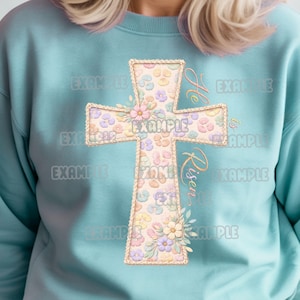 Könnte beinhalten: Hellblaues Sweatshirt mit floralem Kreuz-Design. Das Kreuz ist mit pastellfarbenen Blumen und den Worten "He is Risen" in einer Schriftart verziert. Das Design hat eine Vintage- und Aquarell-Ästhetik.