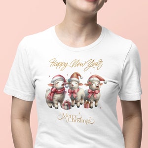 Könnte beinhalten: Weißes T-Shirt mit einem weihnachtlichen Design. Es zeigt drei Cartoon-Schafe mit Weihnachtsmützen und Schals, mit dem Text "Happy New Year" und "Merry Christmas" in goldener Schrift. Die Schafe halten Geschenke.