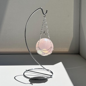 Puede incluir: Una bola de cristal rosa, suspendida por una cadena plateada de un soporte metálico curvo. La esfera está iluminada, mostrando su calidad translúcida. El soporte tiene una base circular, proyectando una sombra sobre la superficie blanca. Decorativo y elegante.