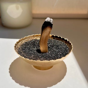 May include: A gold metal bowl with black sand and a smoldering stick of incense. The incense is emitting a thin stream of white smoke.