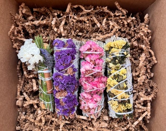 Può includere: Quattro mazzi di erbe e fiori secchi avvolti in salvia bianca, pronti per essere bruciati come incenso. I mazzi sono disposti in una scatola di cartone con carta da imballaggio marrone. I mazzi sono viola, rosa, gialli e bianchi.