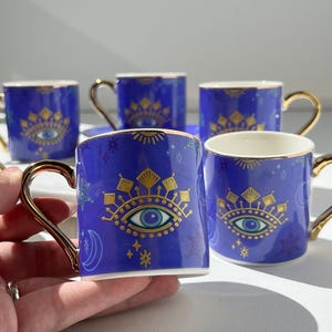 May include: Set of six blue ceramic mugs with gold handles and rims. Each mug features a gold and teal eye design with celestial accents. The mugs are arranged on a white surface, with one mug held in a hand.