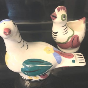May include: A white ceramic dove with green, blue, yellow, and red painted details. The dove is standing next to a white ceramic chicken with black, green, and red painted details. Both figurines have a vintage style.