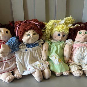 Könnte beinhalten: Vier Vintage-Stoffpuppen mit Garnhaaren und bemalten Gesichtern. Die Puppen tragen Kleider in Rot, Blau, Grün und Rosa. Eine Puppe hat das Wort "TOY" auf ihrem Kleid gestickt. Die Puppen sitzen auf einer grauen Oberfläche.