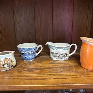 May include: Four ceramic pitchers of varying sizes and designs are displayed on a wooden shelf. The pitchers include a small cream-colored pitcher with a landscape scene, a blue and white patterned pitcher, a pitcher with a landscape scene, and an orange pitcher.