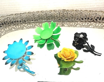 May include: Five vintage flower brooches on a silver tray. The brooches are in the shapes of a green flower, a blue flower, a yellow rose, and two black roses.