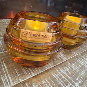 Puede incluir: Dos candelabros de vidrio color ámbar con un diseño en espiral. Cada uno contiene una vela amarilla. El logotipo de Nachtmann y el texto "Hand Washing Recommended" son visibles. Los candelabros están sobre una superficie de madera.