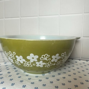 Puede incluir: Un tazón mezclador redondo de color verde oliva con un patrón floral blanco en el exterior. El tazón cuenta con dos pequeñas asas integradas en los lados opuestos y un acabado liso y brillante. El interior es blanco y la base es circular.