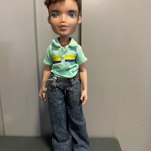Könnte beinhalten: Ein grün-gelb gestreiftes Hemd mit blauem Kragen und einer Puppe mit braunen Haaren, die blaue Jeans und grüne Schuhe trägt.