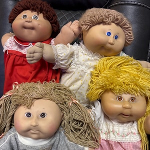 Puede incluir: Cuatro muñecas Cabbage Patch Kids, cada una con un color de pelo y un atuendo diferente. Las muñecas están sentadas en una silla de cuero marrón. Las muñecas llevan un vestido rojo, un vestido blanco con flores amarillas, una sudadera gris y un vestido rosa con flores blancas.