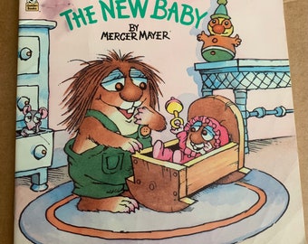 May include: A children's book cover featuring a brown furry creature with a green shirt holding a baby in a wooden cradle. The book title is "The New Baby" by Mercer Mayer.
