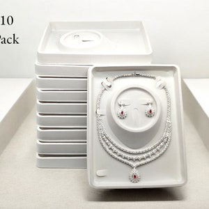 May include: A white jewelry display set with a necklace and earrings. The necklace and earrings are silver-toned with clear stones and red accents. The display set is in a white tray, with a stack of similar trays behind it. The text "10 Pack" is in the upper left corner.