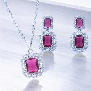 May include: A silver necklace and earrings set with a large, rectangular, dark pink gemstone surrounded by smaller, clear gemstones.