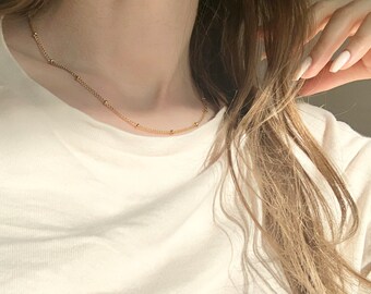 May include: A simple gold chain necklace with small round beads. The necklace is worn by a person with long brown hair.