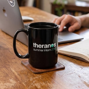 Może przedstawiać: Czarny ceramiczny kubek z białym napisem "theranos", turkusową kropką nad literą "o" oraz nadrukowanym poniżej tekstem "Summer intern 2015". Kubek posiada wygięte ucho i stoi na kwadratowej, brązowej skórzanej podstawce na drewnianym biurku.