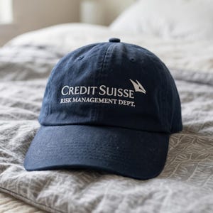 May include: A navy blue baseball cap with the words "Credit Suisse Risk Management Dept." in white. The cap is made of a soft material and has a curved brim. The cap is resting on a gray patterned bedspread.