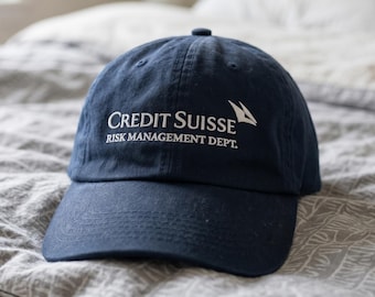 May include: A navy blue baseball cap with the words "Credit Suisse Risk Management Dept." in white. The cap is made of a soft material and has a curved brim. The cap is resting on a gray patterned bedspread.