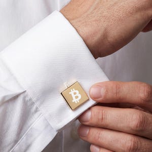 May include: A square gold-toned cufflink featuring a white Bitcoin symbol centered on the face. The cufflink is secured through the buttonholes of a crisp white long-sleeved shirt cuff, showcasing a polished metallic finish and a clean design.