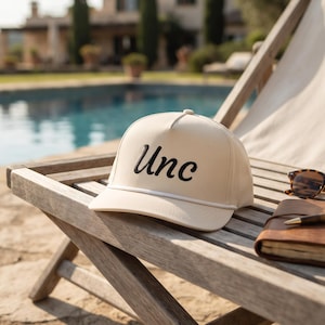 May include: A cream-colored baseball cap featuring the word Unc embroidered in black script on the front panel. The cap includes a white cord detail across the base of the crown and a structured brim, presented alongside a notebook and glasses.