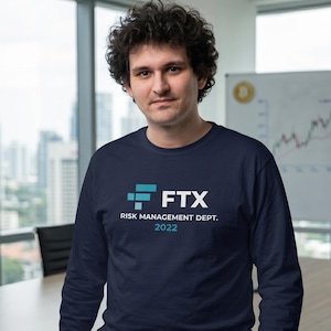 May include: Navy blue long-sleeve shirt with the FTX logo and the text "RISK MANAGEMENT DEPT. 2022" in white. The shirt features a teal and white logo. The person is standing in front of a window.