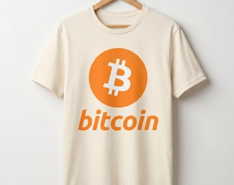 May include: A cream-colored short-sleeve t-shirt featuring a large orange circle with a white Bitcoin symbol in the center. Below the circle, the word bitcoin is printed in orange lowercase letters. The shirt is displayed on a wooden hanger.