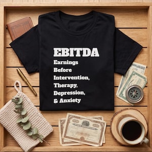 May include: A black short-sleeved t-shirt featuring white text that reads EBITDA Earnings Before Intervention, Therapy, Depression, & Anxiety. The shirt is displayed on a wooden surface alongside a notebook, a pen, currency, and a coffee mug.
