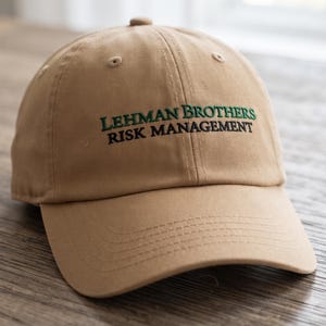 Könnte beinhalten: Eine beigefarbene Baseballkappe mit gesticktem Text auf der Vorderseite. Die Wörter LEHMAN BROTHERS sind in Grün gestickt, während die Wörter RISK MANAGEMENT direkt darunter in Schwarz gestickt sind. Die Kappe hat einen gebogenen Schirm.