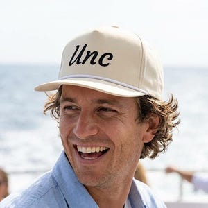 May include: A cream-colored baseball cap featuring the word Unc embroidered in black script font on the front panel. The cap includes a white cord detail across the base of the crown and a structured brim, designed with a classic six-panel construction.