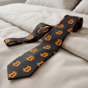 May include: A black necktie featuring a repeating pattern of orange Bitcoin symbols. The tie is laid out flat, displaying a pointed tip and a folded section, showcasing the consistent graphic print across the dark fabric surface.