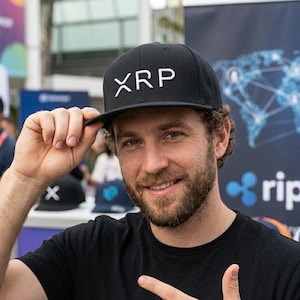 May include: Black baseball cap with the white letters "XRP" on the front. The person in the image is wearing a black t-shirt and pointing at the cap. The background includes a banner with the word "ripple".