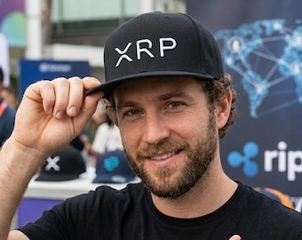 May include: Black baseball cap with the white letters "XRP" on the front. The person in the image is wearing a black t-shirt and pointing at the cap. The background includes a banner with the word "ripple".