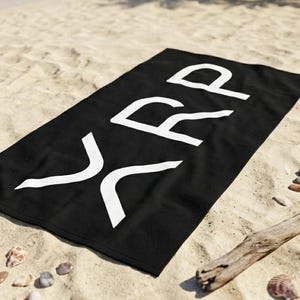 May include: Black beach towel with the white letters "XRP" printed on it. The towel is rectangular and lies on a sandy beach with small rocks, seashells, and driftwood scattered around it. The ocean is visible in the background.