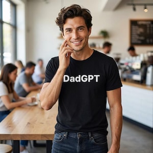 May include: A black t-shirt with the white text "DadGPT" is worn by a person. The person is smiling and touching their face. The background is a cafe setting with other people and a wooden table.