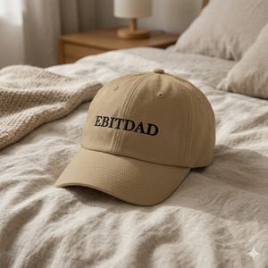 May include: A beige baseball cap featuring the text "EBITDAD" in black lettering. The cap has a curved peak and is made of a soft fabric. It rests on a bed with a neutral-toned duvet cover.