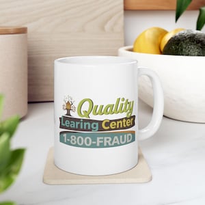 Könnte beinhalten: Weiße Keramiktasse mit den Worten "Quality Learning Center" und "1-800-FRAUD" in Grün, Braun und Blau aufgedruckt. Die Tasse steht auf einem beigen Untersetzer.