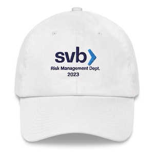 May include: A white baseball cap with the text "svb" in navy blue, followed by a blue arrow. Below the logo is "Risk Management Dept. 2023" in navy blue. The cap has a curved brim and a button on top.