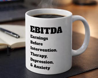May include: White ceramic coffee mug with the text "EBITDA Earnings Before Intervention, Therapy, Depression, & Anxiety" in black. The mug is filled with dark coffee, and steam rises from the top. The mug sits on a dark surface.