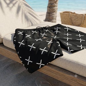 Peut inclure: Short de bain noir avec un motif répétitif en forme de X blanc. Le short est doté d'une taille élastique avec un cordon de serrage noir. Le tissu présente une texture lisse et le motif est réparti uniformément sur le devant du vêtement.
